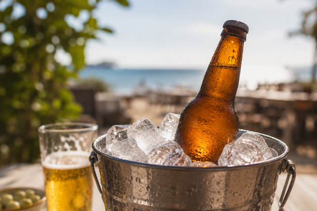Botella de cerveza muy fría en un cubo metálico con hielo, en una terraza junto al mar durante un día de verano.
