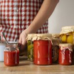 Persona junto a varios tarros de tomate, verduras y setas preparados como conservas caseras.