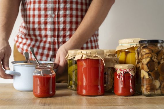 Persona junto a varios tarros de tomate, verduras y setas preparados como conservas caseras.