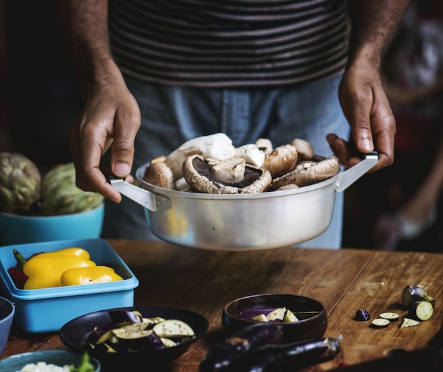 Persona sosteniendo una cazuela con setas junto a verduras sobre una mesa de cocina
